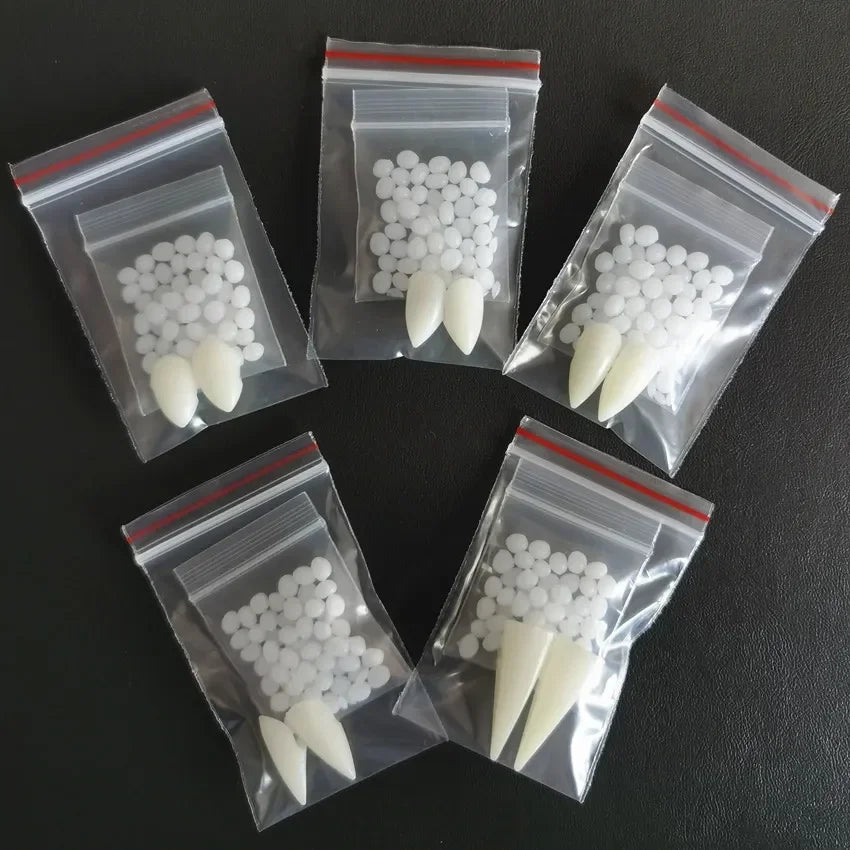A pair of white resin vampire fangs with adhesive pellets in a clear resealable bag, shown against a dark background.